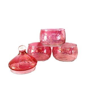 May include: Four pieces of pink glass serveware. There are three small, round bowls and one lidded jar. The glass has a subtle iridescent sheen and a delicate etched leaf design. The pieces are arranged on a white background.