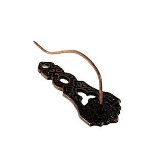 May include: A dark bronze-toned metal wall hook featuring an ornate, elongated backplate with intricate cut-out patterns and a single, curved, slender hook extending outward from the center of the decorative base plate.