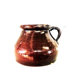 May include: A brown ceramic jar with a handle. The jar has a rounded body and a slightly flared rim. The handle is a simple loop shape. The jar is glazed with a glossy finish.