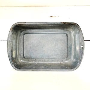 Puede incluir: Una bandeja rectangular para hornear de estilo vintage con bordes redondeados y asas. La bandeja tiene un acabado de esmalte gris moteado y un pequeño agujero en un lado. La bandeja está vacía y se encuentra sobre una superficie blanca.