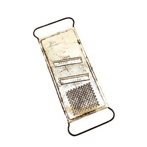 May include: A rectangular metal kitchen grater with a weathered, off-white finish and dark wire handles at both ends. The surface features two horizontal slicing blades and a section with small, raised circular perforations for grating food items.