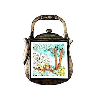 May include: A vintage metal teapot-shaped decorative item. The teapot has a handle and a square tile insert featuring a landscape scene with white daisies, yellow flowers, trees, and a small house. The metal has a tarnished, antique appearance.
