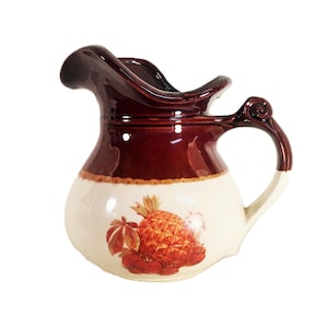 May include: A vintage ceramic pitcher with a bulbous base and a dark brown rim and handle. The pitcher features a cream-colored body with a detailed illustration of a pineapple and leaves in shades of orange and red.