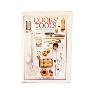 Op de afbeelding: Een kookboek getiteld "COOKS' TOOLS" met diverse keukengerei rondom de titel. De omslag toont een rasp, een lepel, een snijmachine, gember, een muffinvorm en ander kookgerei. De naam van de auteur, SUSAN CAMPBELL, staat onderaan.