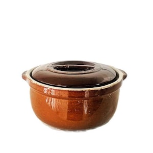 Op de afbeelding: Een ronde, geglazuurde keramische braadpan met een donkerbruin deksel en een lichtere, warmbruine basis. De pan is voorzien van twee kleine, geïntegreerde handgrepen aan de zijkanten en heeft een gladde, glanzende afwerking met een functioneel ontwerp.