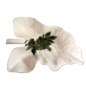 May include: A white ceramic dish shaped like a large leaf with a textured surface. The center of the dish features a raised, green-glazed cluster of grapes with detailed leaves, and a small stem extends from the left side of the leaf-shaped bowl.