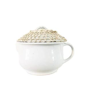 May include: A white ceramic bowl with a handle and a matching lid. The lid is covered with a beige, open-work crocheted fabric overlay that features a net-like pattern. The bowl has a smooth, glossy finish and a rounded, wide-bottomed shape.