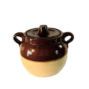 Pode incluir: Um pote de cerâmica de dois tons com uma seção superior esmaltada em castanho escuro brilhante e uma seção inferior em bege mate. O recipiente possui duas alças laterais e uma tampa removível a condizer com um puxador central.