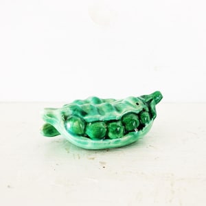 Puede incluir: Figura de vaina de guisantes de cerámica en tonos verdes. La vaina está abierta, revelando varios guisantes redondos. La figura tiene un acabado brillante y está sobre un fondo blanco. Un artículo decorativo.