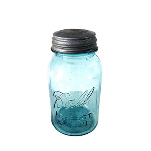Op de afbeelding: Een vintage lichtblauwe glazen mason jar met een metalen schroefdeksel. De voorzijde van de pot is voorzien van de in reliëf aangebrachte tekst Ball in cursief schrift en de tekst PERFECT MASON in blokletters. Het glas heeft een aqua tint.