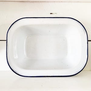 May include: White enamelware rectangular baking dish with a dark blue rim. The dish has rounded corners and a slightly raised edge. The background is a white wooden surface.