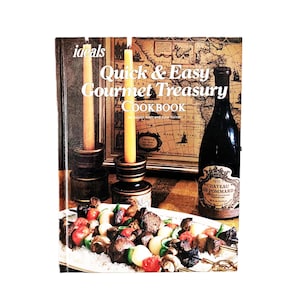 以下が含まれることがあります： 「Ideals Quick & Easy Gourmet Treasury Cookbook」というタイトルのハードカバー料理本。表紙にはヴィンテージ風の地図、暗い色の燭台に立てられた2本の黄色いテーパーキャンドル、暗い色のワインボトル、そして肉と野菜の串焼きが盛り付けられた大皿が描かれています。