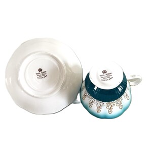 May include: A white porcelain teacup and saucer set featuring a teal band and gold scrollwork patterns. The underside of both pieces displays the Royal Albert Bone China England Overture Series stamp with a crown logo in black text.