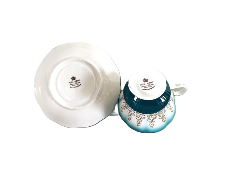 May include: A white porcelain teacup and saucer set featuring a teal band and gold scrollwork patterns. The underside of both pieces displays the Royal Albert Bone China England Overture Series stamp with a crown logo in black text.
