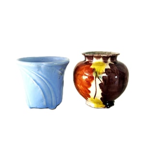 Könnte beinhalten: Ein hellblauer Keramikübertopf mit ausgestelltem Rand und geprägten geschwungenen Linien neben einer kleinen, runden Keramikvase mit glänzender, mehrfarbiger Glasur in Orange, Gelb und Dunkelbraun mit einer breiten, kreisförmigen Öffnung.