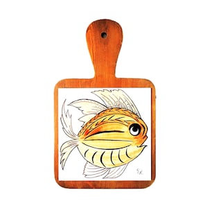 May include: Wooden chopping board with a handle, featuring a hand-painted fish design. The fish is yellow, orange, and black, set against a white tile background. The board is made of wood and has a natural finish.