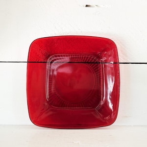 May include: A square, ruby-red glass plate with a textured surface and a slightly raised centre. The plate has rounded corners and a black trim. The plate is sitting on a white surface.