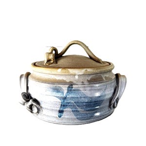 Op de afbeelding: Een keramische pot met deksel en twee zijhandvatten, voorzien van een lichtgrijze basis met blauwe penseelstreekpatronen. Het deksel heeft een gebogen handvat en een klein decoratief sculpturaal element. De rand heeft een bruine glazuur.