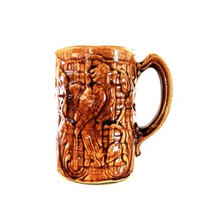 May include: A brown ceramic mug featuring a raised relief design of a bird perched on a branch. The exterior surface has a glossy finish with a textured, grid-like pattern surrounding the central bird motif and a curved handle on the right side.