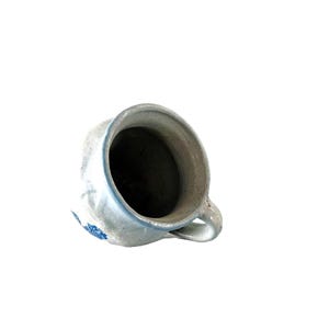 May include: A rustic, off-white ceramic pitcher with a handle and a blue floral design. The pitcher has a slightly distressed appearance, suggesting it is vintage or handmade. The interior is dark, contrasting with the light exterior.