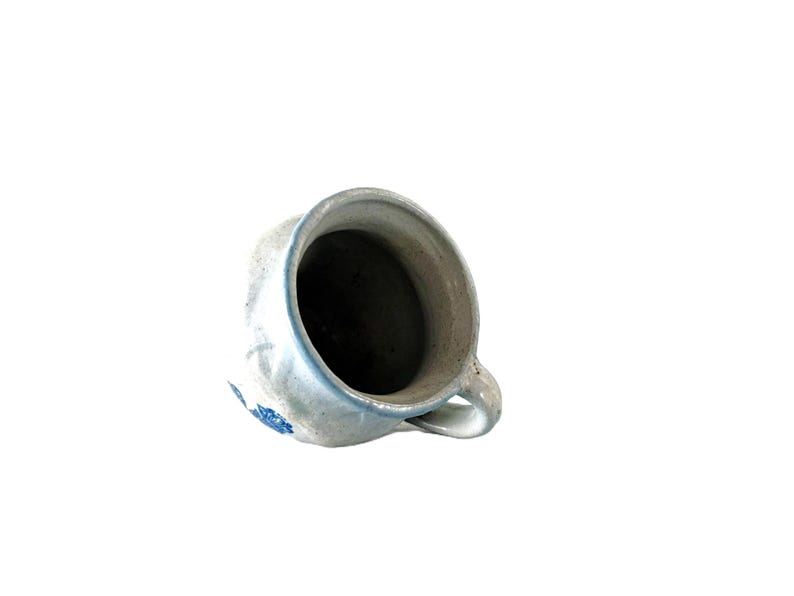 May include: A rustic, off-white ceramic pitcher with a handle and a blue floral design. The pitcher has a slightly distressed appearance, suggesting it is vintage or handmade. The interior is dark, contrasting with the light exterior.