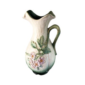 May include: A ceramic pitcher featuring a cream-colored body with a textured, bark-like surface. The vessel is adorned with raised, sculpted green leaves and light purple flowers with yellow centers. It has a curved handle and a scalloped rim.