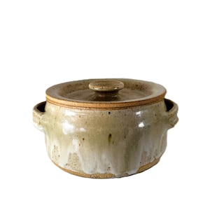 Puede incluir: Una olla de cerámica redonda con tapa y dos asas laterales, con un esmalte moteado en tonos beige y bronceado que se transforma en un patrón de goteo blanco hacia la base. La olla tiene una tapa redondeada y lisa con un pequeño pomo central.