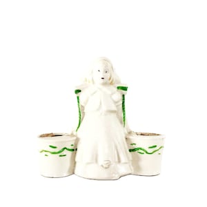 May include: A vintage white ceramic planter in the shape of a woman, with two side planters. The woman figure has green accents on her shoulders and the planters have green wave designs. A unique decorative piece.
