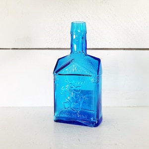 Puede incluir: Una botella de vidrio azul brillante con un cuerpo rectangular y una parte superior en forma de casa. La botella presenta un diseño en relieve de un caballo y un jinete. La botella está vacía y se encuentra sobre una superficie blanca.