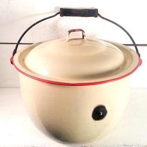 Może przedstawiać: Kremowy garnek emaliowany z pokrywką i czerwoną obwódką. Posiada czarny uchwyt i czarny uchwyt na pokrywce. Garnek ma czarną plamę z boku. Zabytkowy artykuł kuchenny.