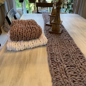 May include: A brown and white chunky knit table runner on a whitewashed wooden table. The runner is made of thick yarn and has a braided texture.