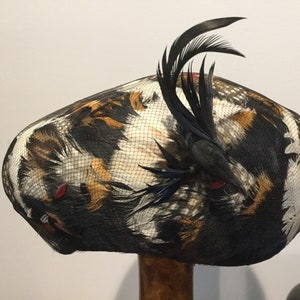 May include: A black, white, and brown feathered hat with a black feather accent. The hat is made of a mesh material and has a round shape.