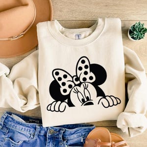 Op de afbeelding: Cr&egrave;mekleurige sweatshirt met een zwart-wit Minnie Mouse-afbeelding. Het ontwerp bevat Minnie's iconische strik met stippen en haar gezicht. De sweatshirt wordt gecombineerd met een blauwe spijkerbroek, een beige hoed en sandalen.