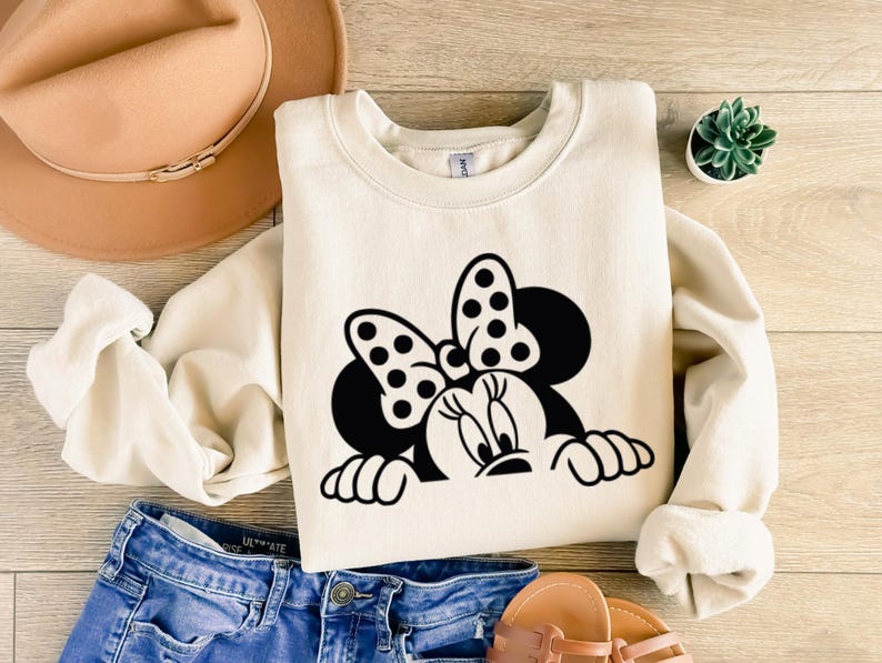 Op de afbeelding: Cr&egrave;mekleurige sweatshirt met een zwart-wit Minnie Mouse-afbeelding. Het ontwerp bevat Minnie's iconische strik met stippen en haar gezicht. De sweatshirt wordt gecombineerd met een blauwe spijkerbroek, een beige hoed en sandalen.