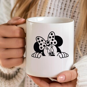 Op de afbeelding: Witte keramische mok met een zwart-wit Minnie Mouse-ontwerp. Het ontwerp bevat Minnie's iconische stippen strik en lachende gezicht. De mok wordt vastgehouden door een persoon die een witte trui draagt.