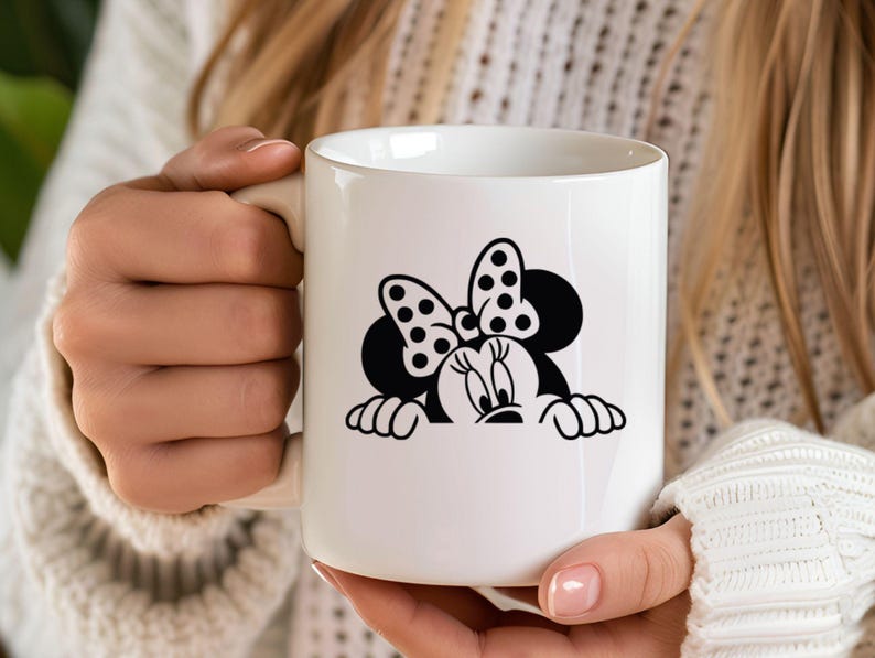Op de afbeelding: Witte keramische mok met een zwart-wit Minnie Mouse-ontwerp. Het ontwerp bevat Minnie's iconische stippen strik en lachende gezicht. De mok wordt vastgehouden door een persoon die een witte trui draagt.