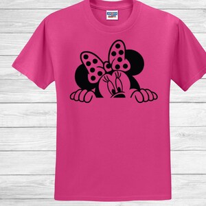 Op de afbeelding: Een felroze t-shirt met een zwarte afbeelding van een stripfiguur met een stippenstrik. Het gezicht en de handen van het personage zijn zichtbaar en kijken over de onderkant van het shirt. Het shirt is gemaakt van een zacht materiaal.