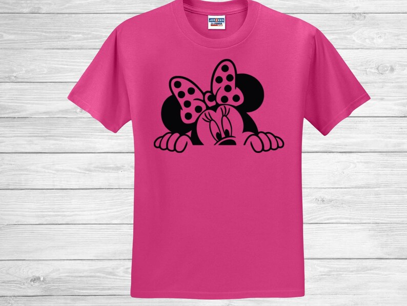 Op de afbeelding: Een felroze t-shirt met een zwarte afbeelding van een stripfiguur met een stippenstrik. Het gezicht en de handen van het personage zijn zichtbaar en kijken over de onderkant van het shirt. Het shirt is gemaakt van een zacht materiaal.