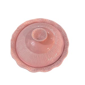 Könnte beinhalten: Eine rosa Keramik-Butterdose mit Deckel. Die Schale hat ein strukturiertes, geriffeltes Muster und einen gewellten Rand. Der Deckel hat einen zentralen Knopf mit einem ähnlichen strukturierten Design. Die Gesamtform ist rund.