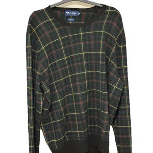 May include: A Polo Golf crew neck sweater in a classic plaid pattern. The sweater features a dark green base with a grid of yellow and red lines. The Polo Golf label is visible on the inside of the collar. The sweater is hanging on a black hanger.
