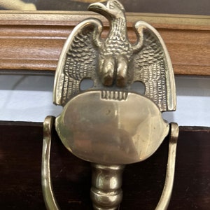 Puede incluir: Un llamador de puerta de latón con un diseño de águila. El águila está posada sobre una placa redonda con un espacio en blanco para grabar. El llamador está unido a un asa curva.