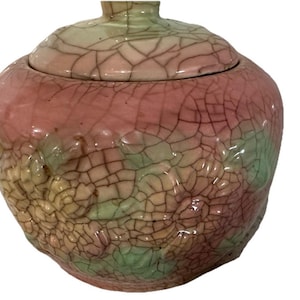 May include: A ceramic jar with a lid, featuring a crackle glaze. The jar is primarily pink, transitioning to green and yellow at the base. The lid is a lighter pink and green. The jar has a rounded shape and a glossy finish.