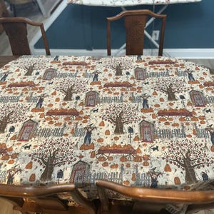May include: A rectangular tablecloth with a cream-colored background featuring a repeating pattern of autumn-themed illustrations including orange pumpkins, brown trees, wooden fences, black cats, and figures wearing hats.