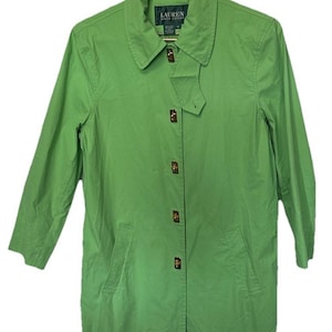 Könnte beinhalten: Ein hellgrüner Trenchcoat mit Kragen und Knopfverschluss. Der Mantel hat zwei Vordertaschen und ein kleines Logo-Etikett in der Nähe des Kragens. Der Mantel hängt an einem Metallbügel.