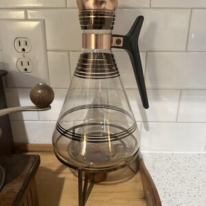 Può includere: Caraffa da caffè vintage in vetro con dettagli color rame e manico nero. La caraffa ha una forma conica con strisce nere orizzontali e poggia su un supporto in metallo. Il coperchio è color rame. Un design classico.