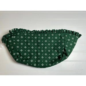 May include: A green fabric valance with a white floral pattern and a ruffled edge. The valance is curved and appears to be made of a soft material. The background is a white wooden surface.