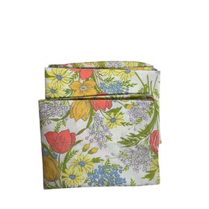 May include: Folded bedding set featuring a floral pattern. The design includes yellow daisies, red tulips, blue forget-me-nots, and green foliage on a white background. The bedding set is folded and stacked, showcasing the full pattern.