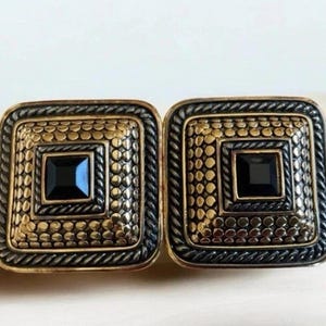 Könnte beinhalten: Zwei quadratische Ohrringe mit einem Gold- und Schwarz-Design. Jeder Ohrring hat einen schwarzen quadratischen Mittelstein, umgeben von einem goldenen Punktmuster und einem schwarzen, seilartigen Rand. Die Ohrringe liegen auf einer hellen Oberfläche.