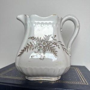 May include: A white ceramic cream jug with a scalloped rim and a curved handle. The body features a brown transferware floral pattern with fern-like leaves and small flowers. The base of the jug has a subtle gold-toned trim detail.