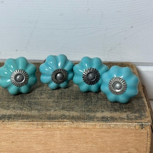May include: Four turquoise ceramic flower-shaped cabinet knobs with silver-colored metal centers. The knobs are arranged in a row on a vintage book. The knobs are a decorative hardware item.
